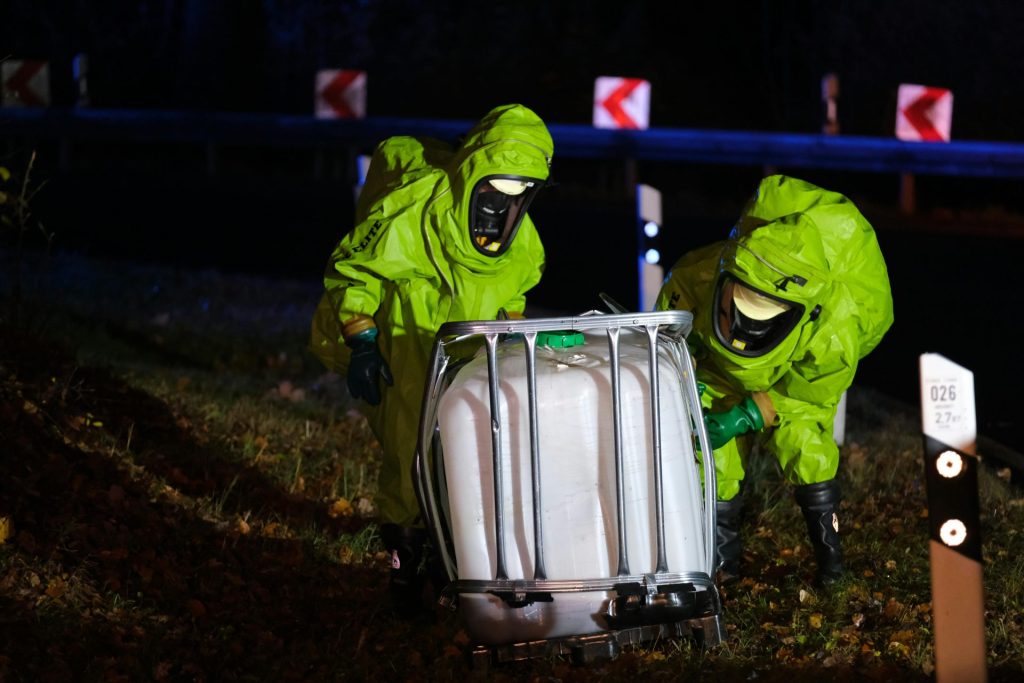 Two firefighters in hazmat suits managing a chemical spill by the roadside at night.
